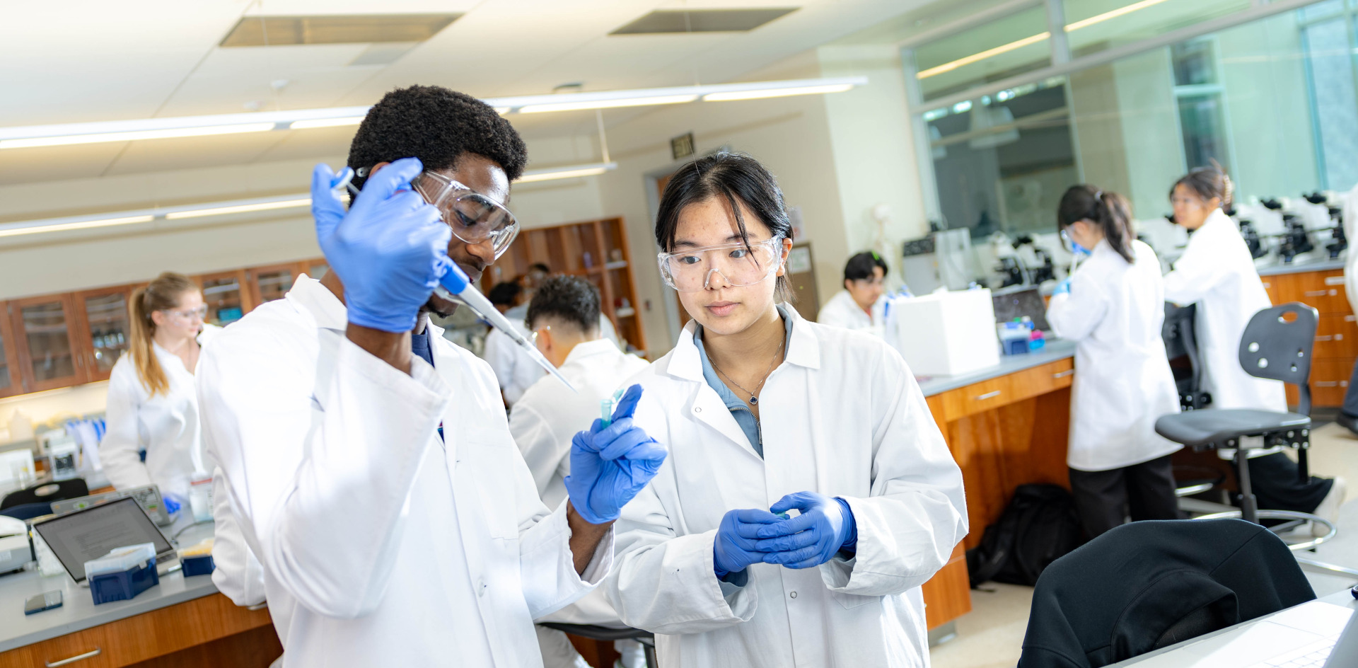 Two students wearing lab coats, blue gloves, and safety glasses perform an experiments using test tubes in a lab.