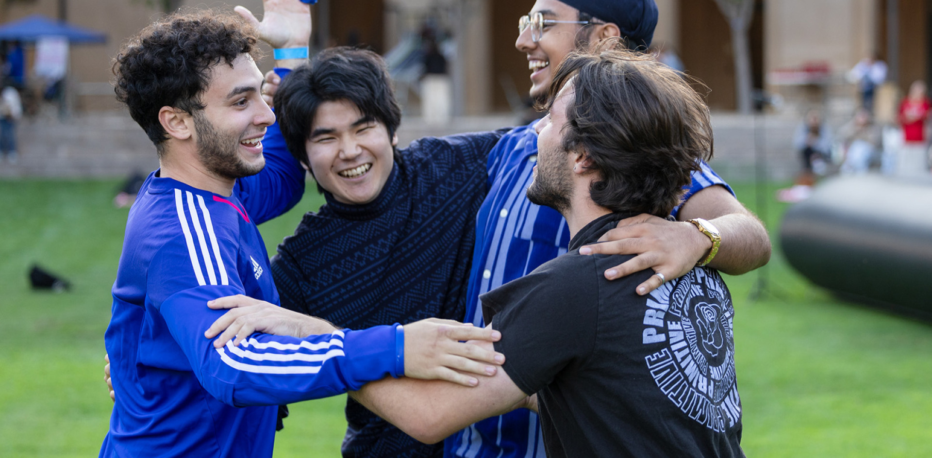  Four young men laughing and hugging on a grassy field.