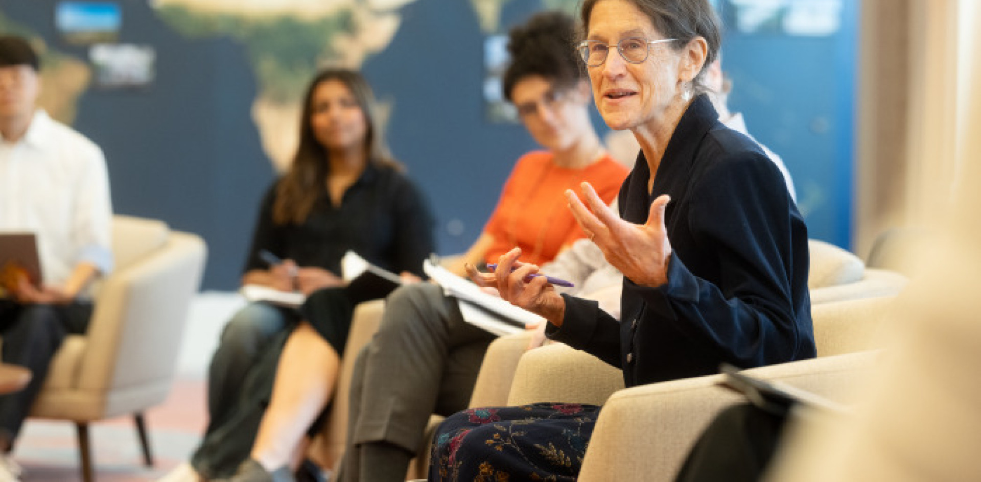 A woman with pulled back brown hair speakers to students looking on as they sit in front of a world map covered wall.