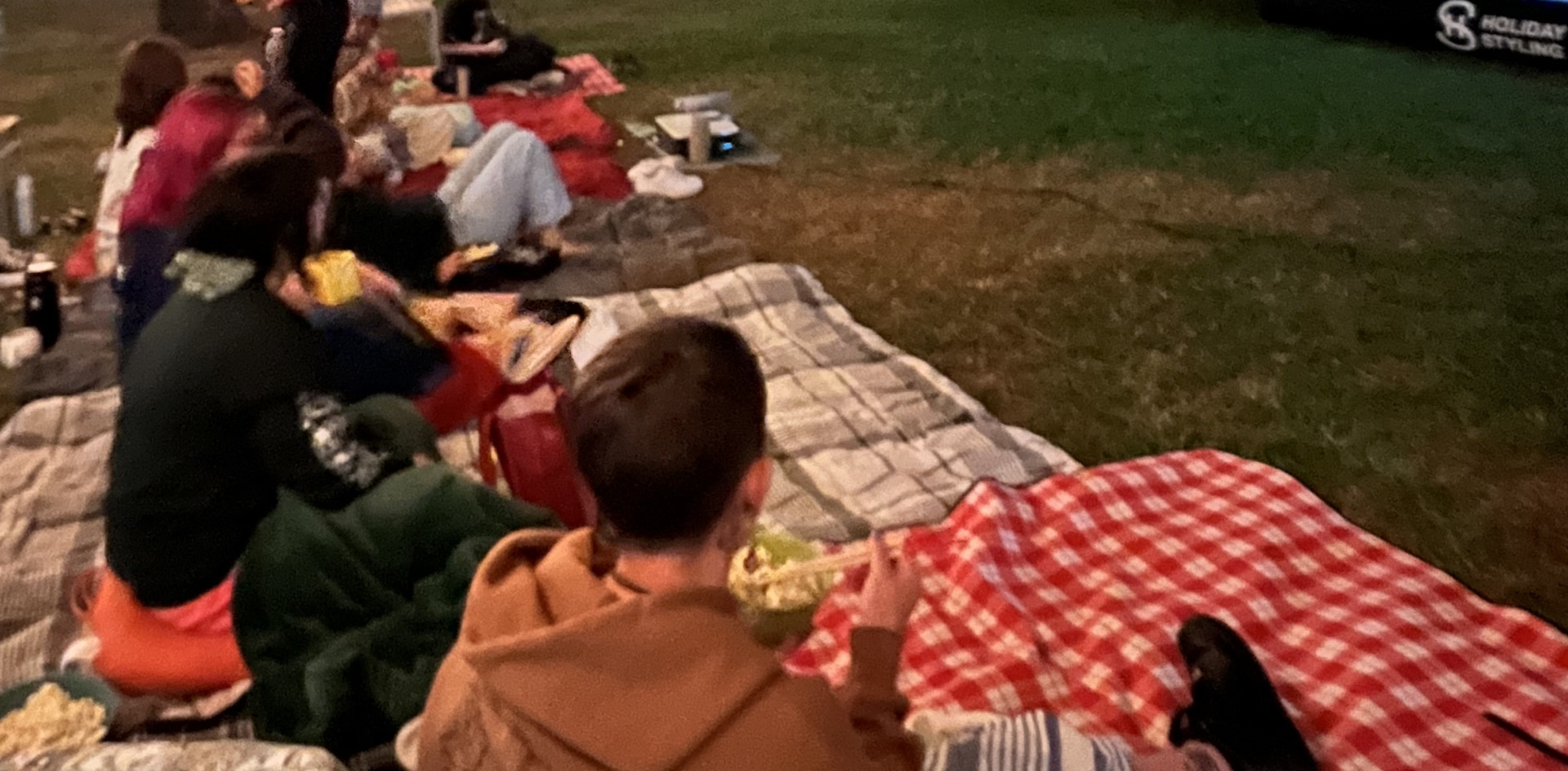 Students sit on blankets under stringlights as they watch a movie on a projected screen outdoors.
