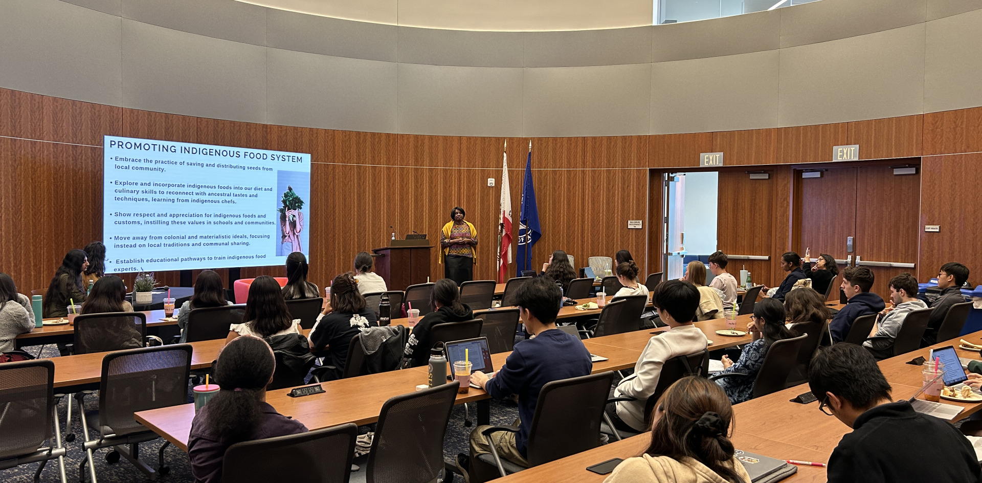 A woman stands next to a lectern and a projector screen as she gives a presentation in an auditorium full of students.