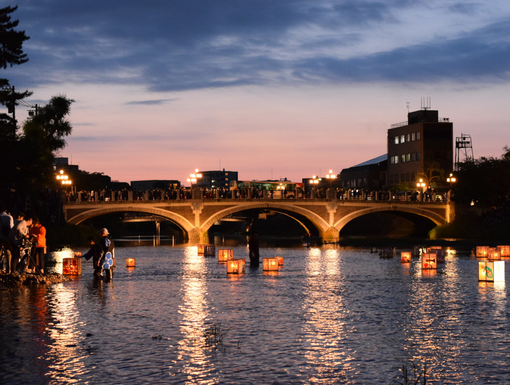 Picture of a bridge illuminated from behind by a sunset and underneath by lanterns floating on the water.
