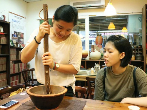 Two women look at a bowl while one of the women stirs with a large wooden stick.
