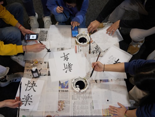 Overhead shot of students gathered around a table practicing Chinese calligraphy.