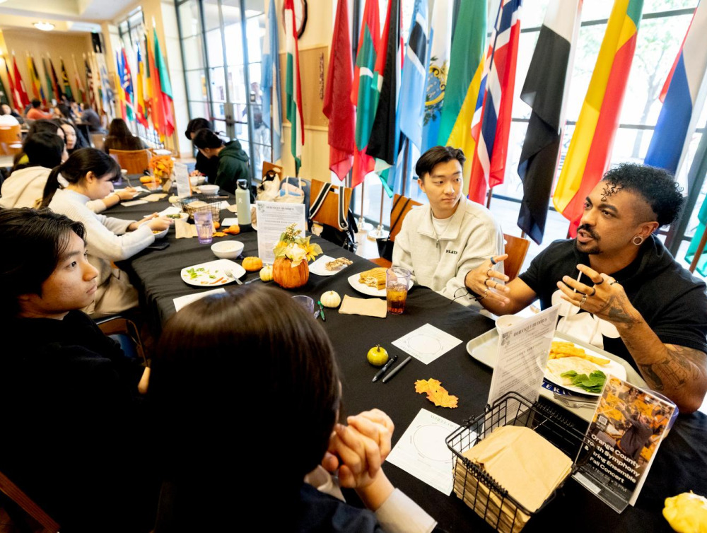 Participants at a "Dialogue Buddies" event engage in conversation around a table, surrounded by international flags and decorated with small pumpkins and flowers.