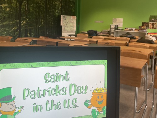 A sign reading "Saint Patricks Day in the US" is displayed at the entrance of a classroom with desks