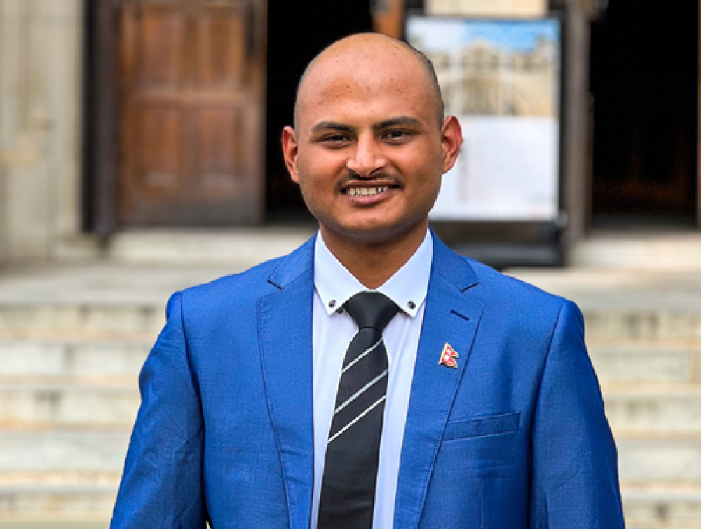 Mukesh Bastola wears a blue suit jacket and striped black tie as he stands in front of marble stairs outside a building with wooden doors.