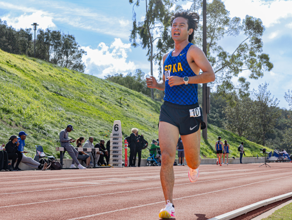 A male with medium length brown hair is wearing a blue checkered tank top with yellow letters spelling SOKA as he runs the track during a track meet.