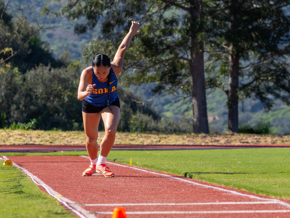 A female track athlete holds her left arm up behind her as she prepares to participate in a long jump competition.