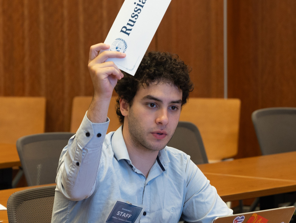 A Soka University Model United Nations team member with curly hair sits at a conference table with a laptop, raising a placard that reads "Russia" while a placard for "Spain" sits on the table in front of him.