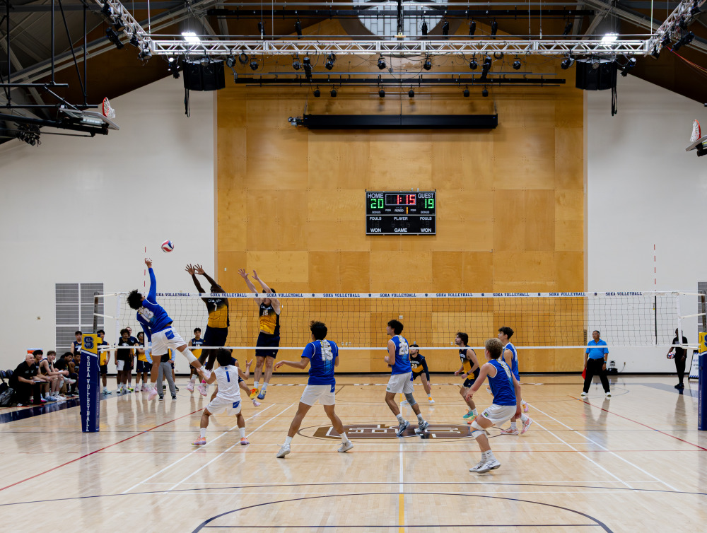 Two teams compete in a men's volleyball match. A SUA player is hitting the ball over the net while the opposing team has two players jumping up trying to block him.