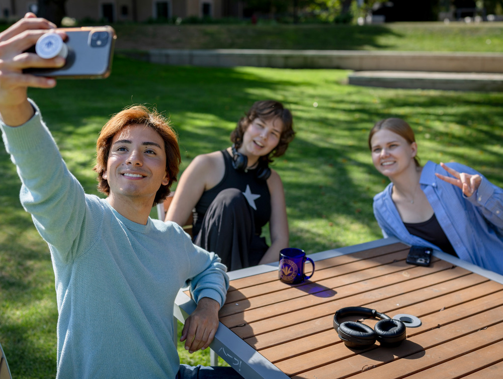  A young man takes a selfie with two friends while sitting at an outdoor wooden table on a sunny campus lawn.