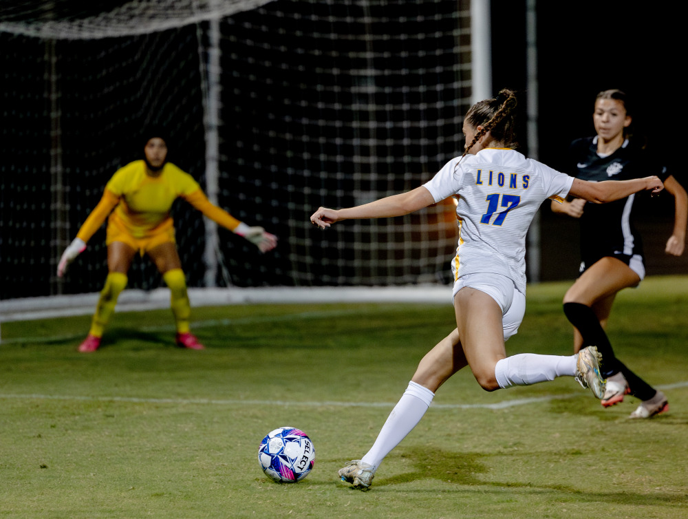 A female soccer player shoots past a defender at a goalkeeper standing in goal.