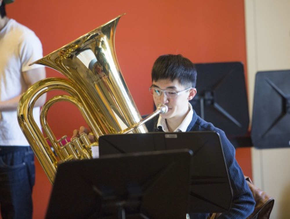 A male student with short black hair and glasses plays a large brass instrument in front of a dark orange wall.