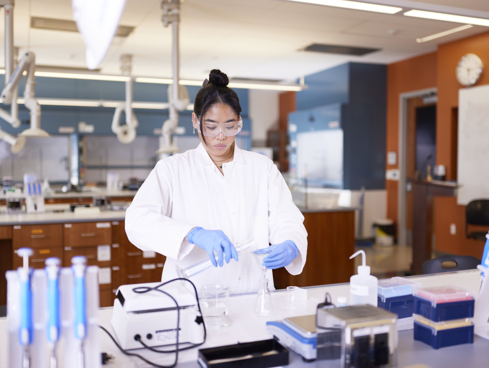 A female student with black tied-back hair wears a white lab coat, safety glasses, and blue gloves as she performs science experiments in a lab.