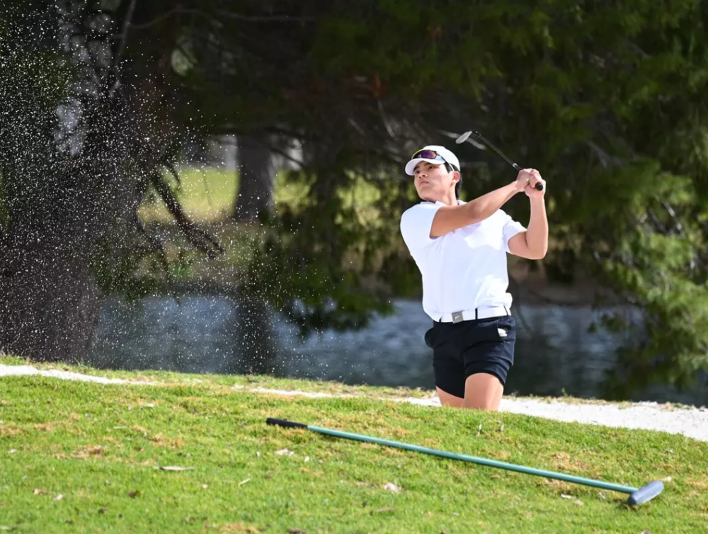 A male golfer wearing a white t-shirt and black shorts watches the ball as he is holding his backswing on a golfcourse.
