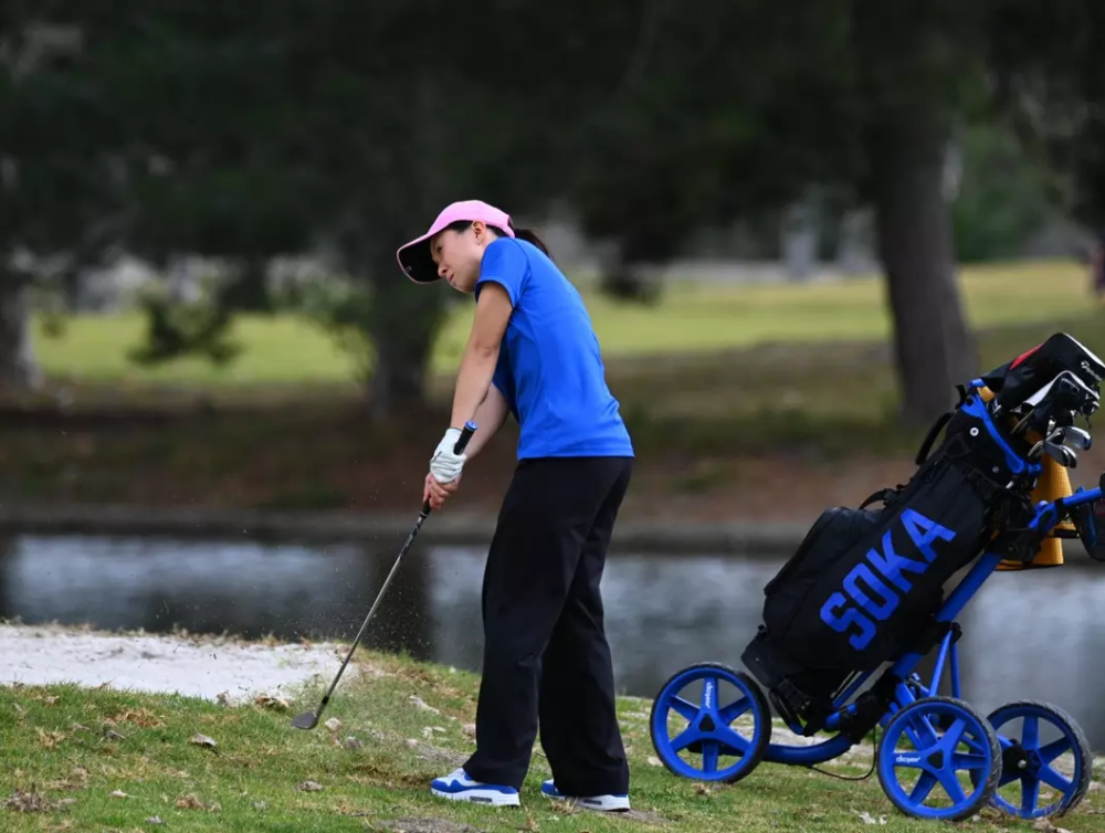A female golfer wearing a blue shirt, black pants, and pink visor hits a golf ball.