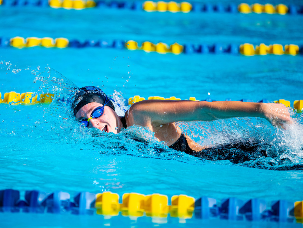 A female swimmer wearing a black swim cap and blue goggles comes up to take a breath during a swim meet.