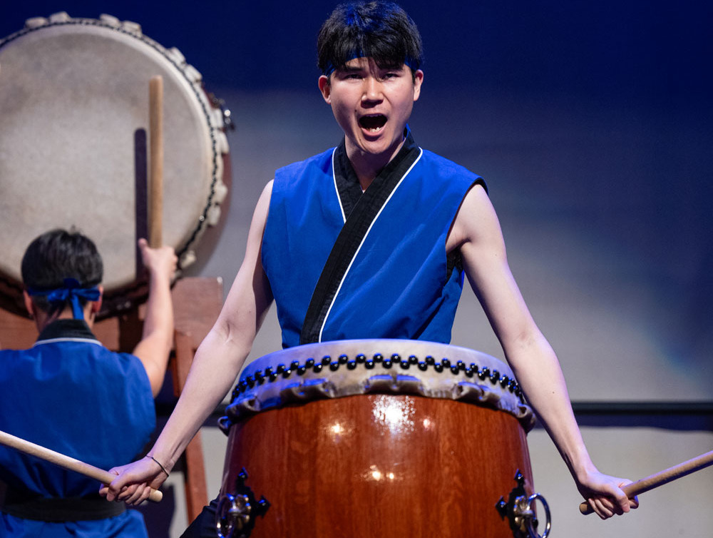 A man with short black hair wearing a royal blue sleeveless shirt with black and white lining stands in front of a large wooden drum holding two drumsticks as he yells during a performance.