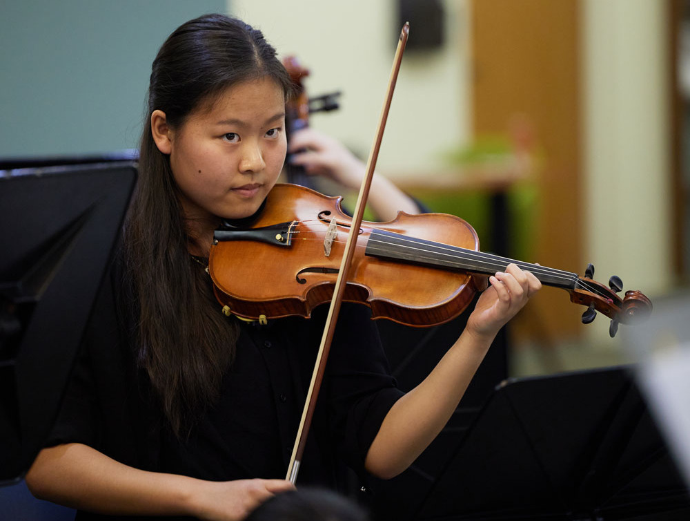 A woman with long dark brown hair wears a black shirt and plays the violin while looking ahead.