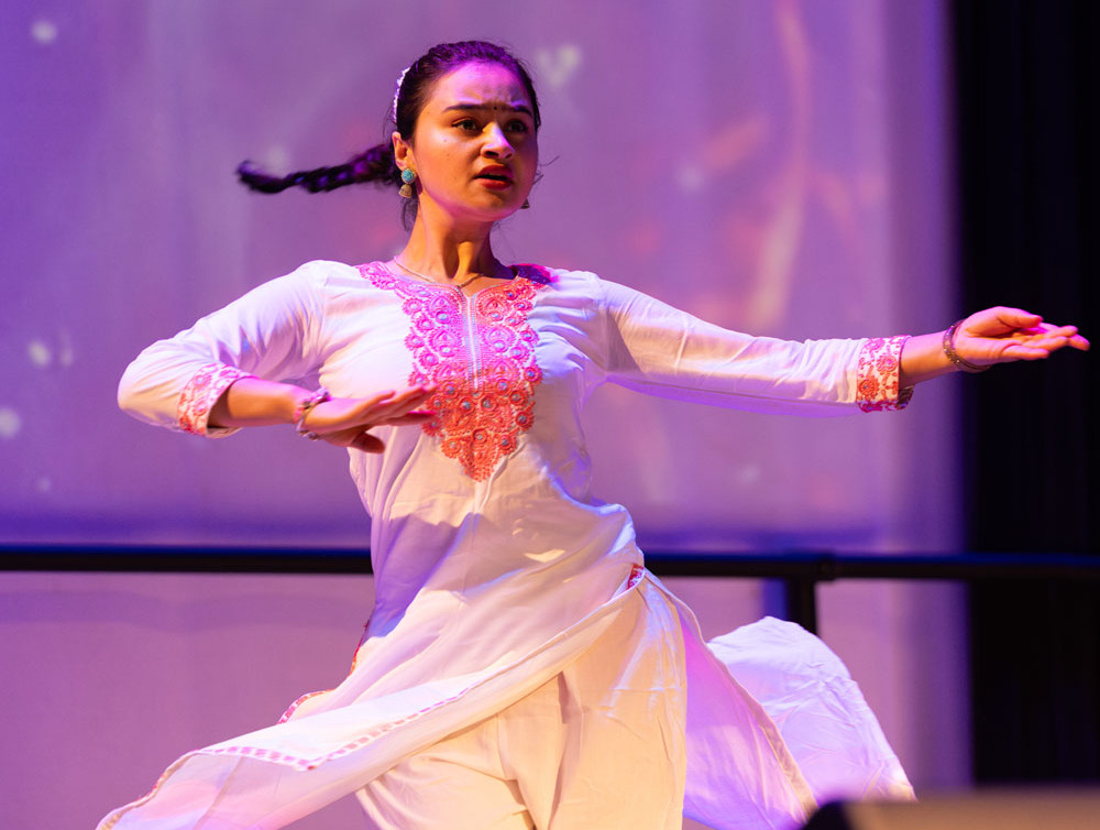 A woman with long, tied-back, brown hair wearing a white and pink dress shirt and pants performs as a Nepali dance.