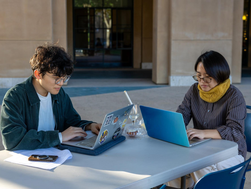 Two students sitting outside the library, working on their laptops