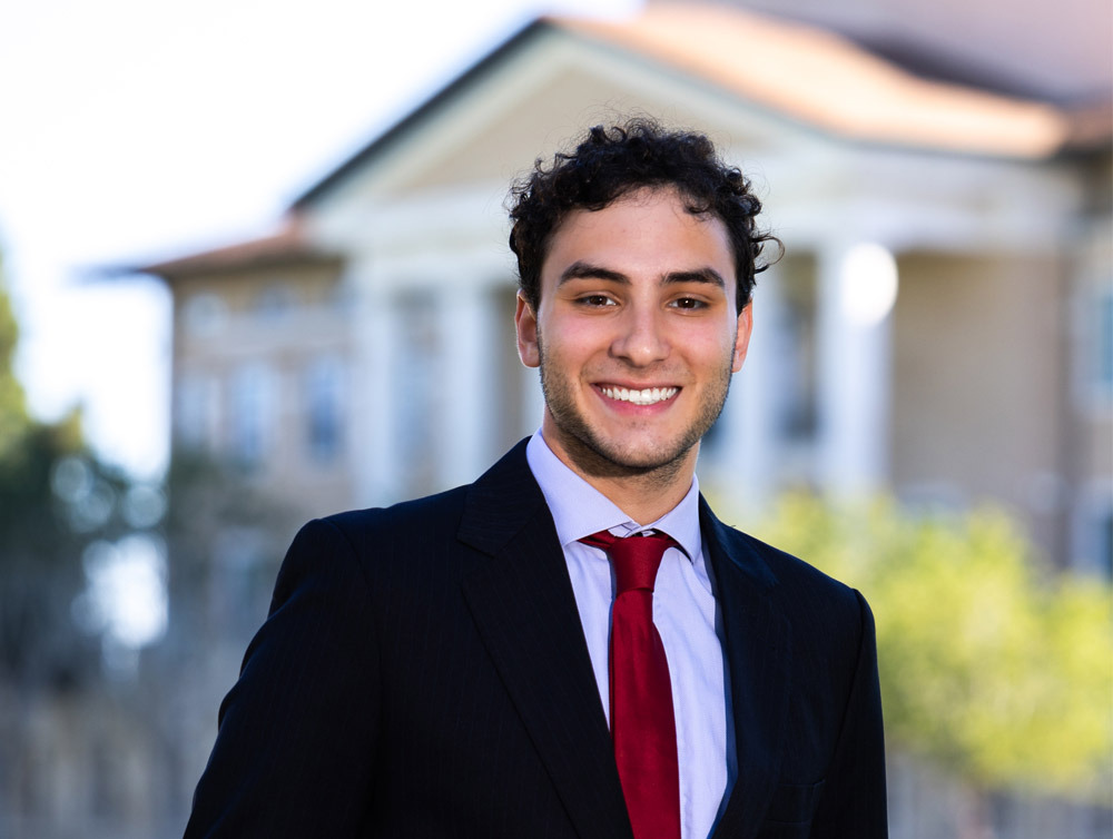 Mateus Quesada wears a black suit jacket over a light blue dress shirt with a red tie as he smiles for a photo outside of Founders Hall.