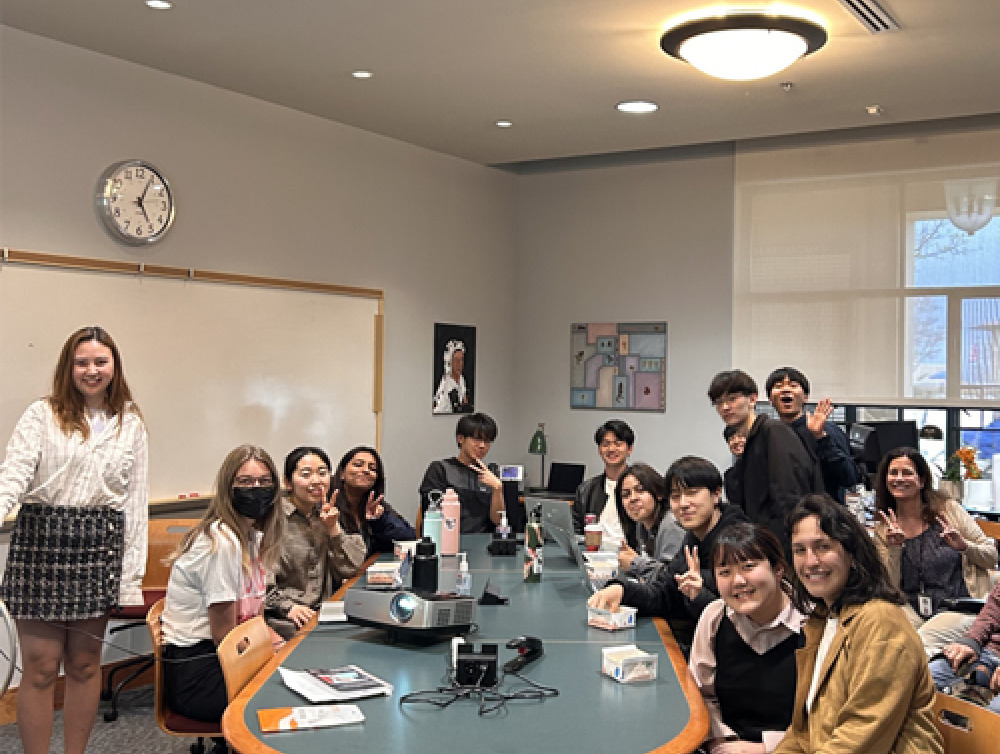 Many students are gathered around a large table smiling at the camera. 