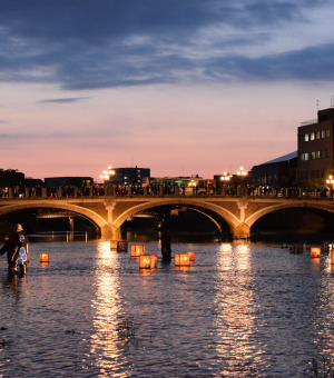 Picture of a bridge illuminated from behind by a sunset and underneath by lanterns floating on the water.