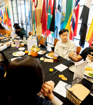 Participants at a "Dialogue Buddies" event engage in conversation around a table, surrounded by international flags and decorated with small pumpkins and flowers.