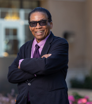 Herbie Hancock wears a black suit jacket over a light purple shirt and a dark purple tie. He is wearing tinted sunglasses and smiles as he stands with his arms crossed outside on Soka's campus.