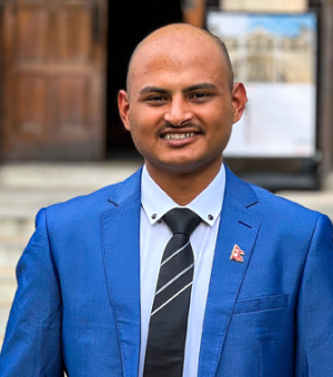 Mukesh Bastola wears a blue suit jacket and striped black tie as he stands in front of marble stairs outside a building with wooden doors.