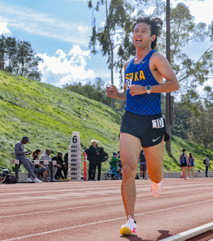 A male with medium length brown hair is wearing a blue checkered tank top with yellow letters spelling SOKA as he runs the track during a track meet.