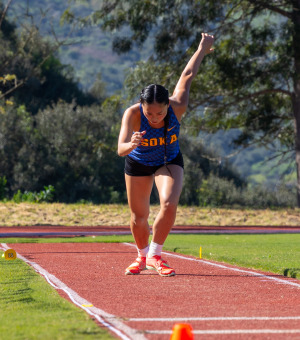 A female track athlete holds her left arm up behind her as she prepares to participate in a long jump competition.