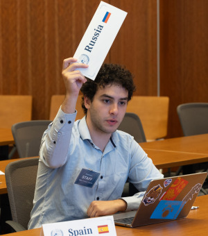 A Soka University Model United Nations team member with curly hair sits at a conference table with a laptop, raising a placard that reads "Russia" while a placard for "Spain" sits on the table in front of him.