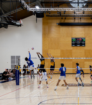 Two teams compete in a men's volleyball match. A SUA player is hitting the ball over the net while the opposing team has two players jumping up trying to block him.