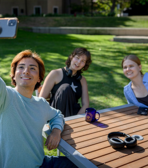  A young man takes a selfie with two friends while sitting at an outdoor wooden table on a sunny campus lawn.