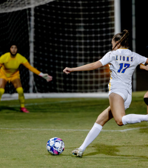 A female soccer player shoots past a defender at a goalkeeper standing in goal.