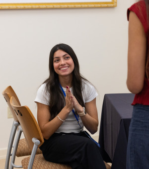 A woman with long brown hair holds her hands together and smiles while looking at people she is conversing with.