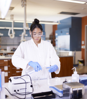 A female student with black tied-back hair wears a white lab coat, safety glasses, and blue gloves as she performs science experiments in a lab.