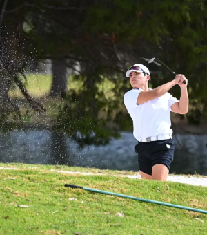 A male golfer wearing a white t-shirt and black shorts watches the ball as he is holding his backswing on a golfcourse.