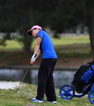 A female golfer wearing a blue shirt, black pants, and pink visor hits a golf ball.