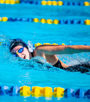 A female swimmer wearing a black swim cap and blue goggles comes up to take a breath during a swim meet.