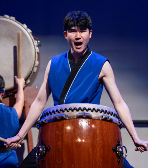 A man with short black hair wearing a royal blue sleeveless shirt with black and white lining stands in front of a large wooden drum holding two drumsticks as he yells during a performance.