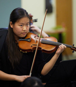 A woman with long dark brown hair wears a black shirt and plays the violin while looking ahead.