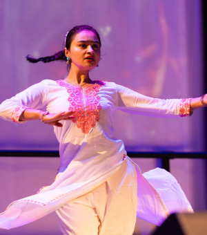 A woman with long, tied-back, brown hair wearing a white and pink dress shirt and pants performs as a Nepali dance.