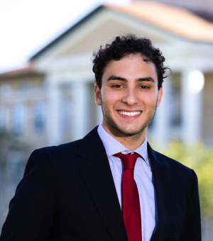 Mateus Quesada wears a black suit jacket over a light blue dress shirt with a red tie as he smiles for a photo outside of Founders Hall.
