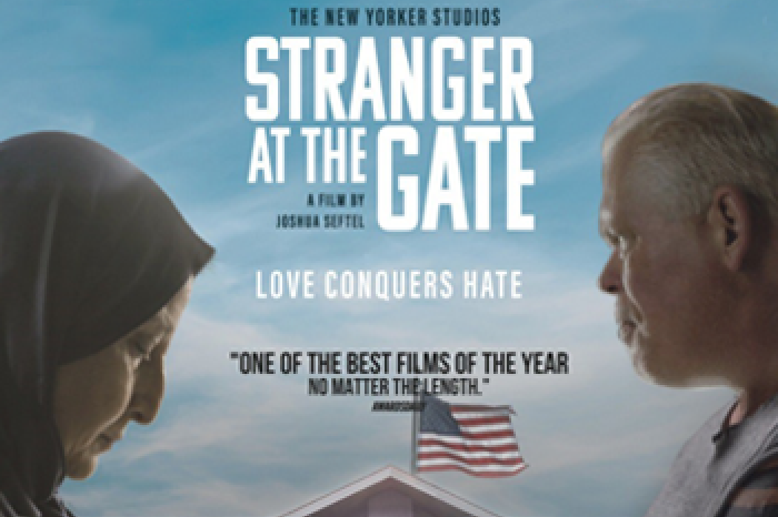 Stranger at the Gate Movie Cover showing two people facing eachother. The person on the left is a woman wearing a headscarf and the person on the right is a white man. They appear to be going head to head over a building with an American flag displayed.