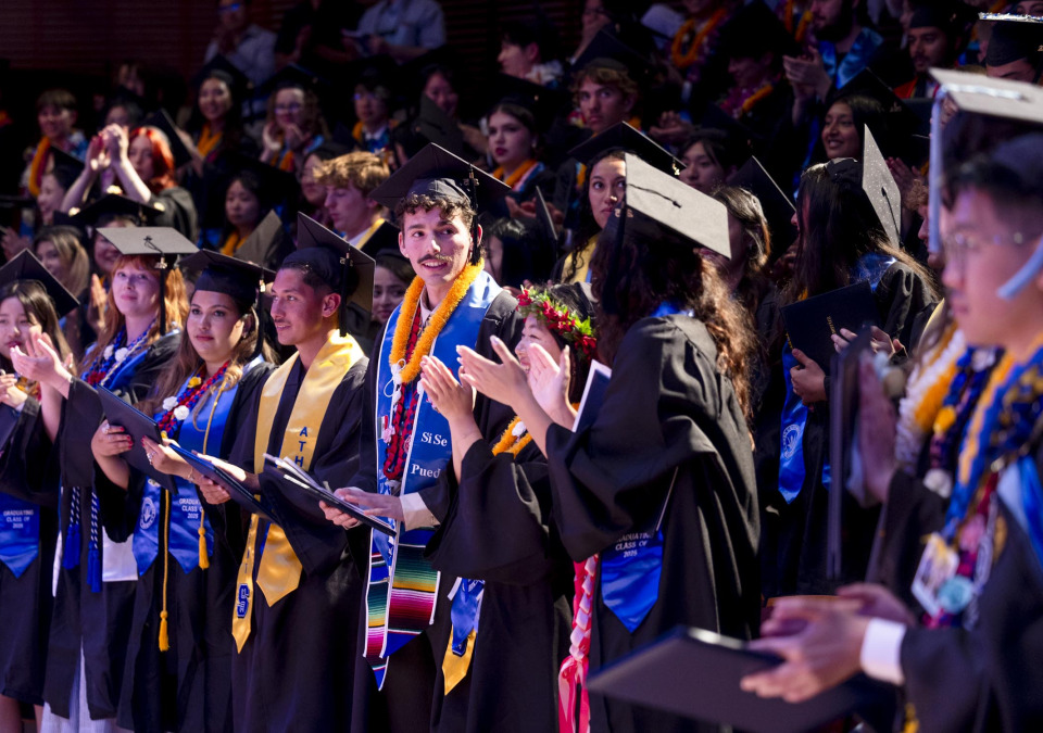 Graduates, wearing their cap and gown, stand and applaud during the 2025 commencement ceremony.