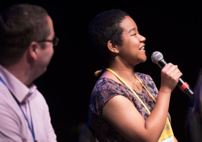 A woman speaks into a microphone while a man listens.